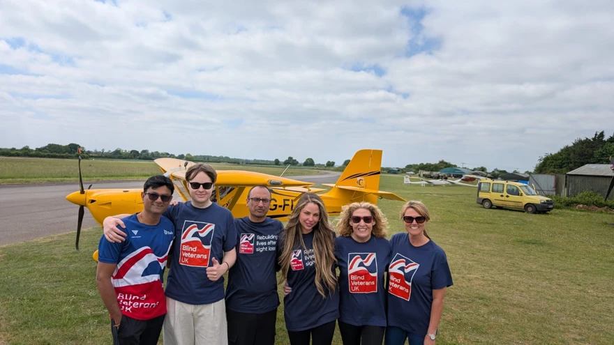 All six of Team Thea stood in front of a small yellow plane which is sat on a grassy field with a runway behind. The team are all wearing Blind Veterans UK T-shirts.