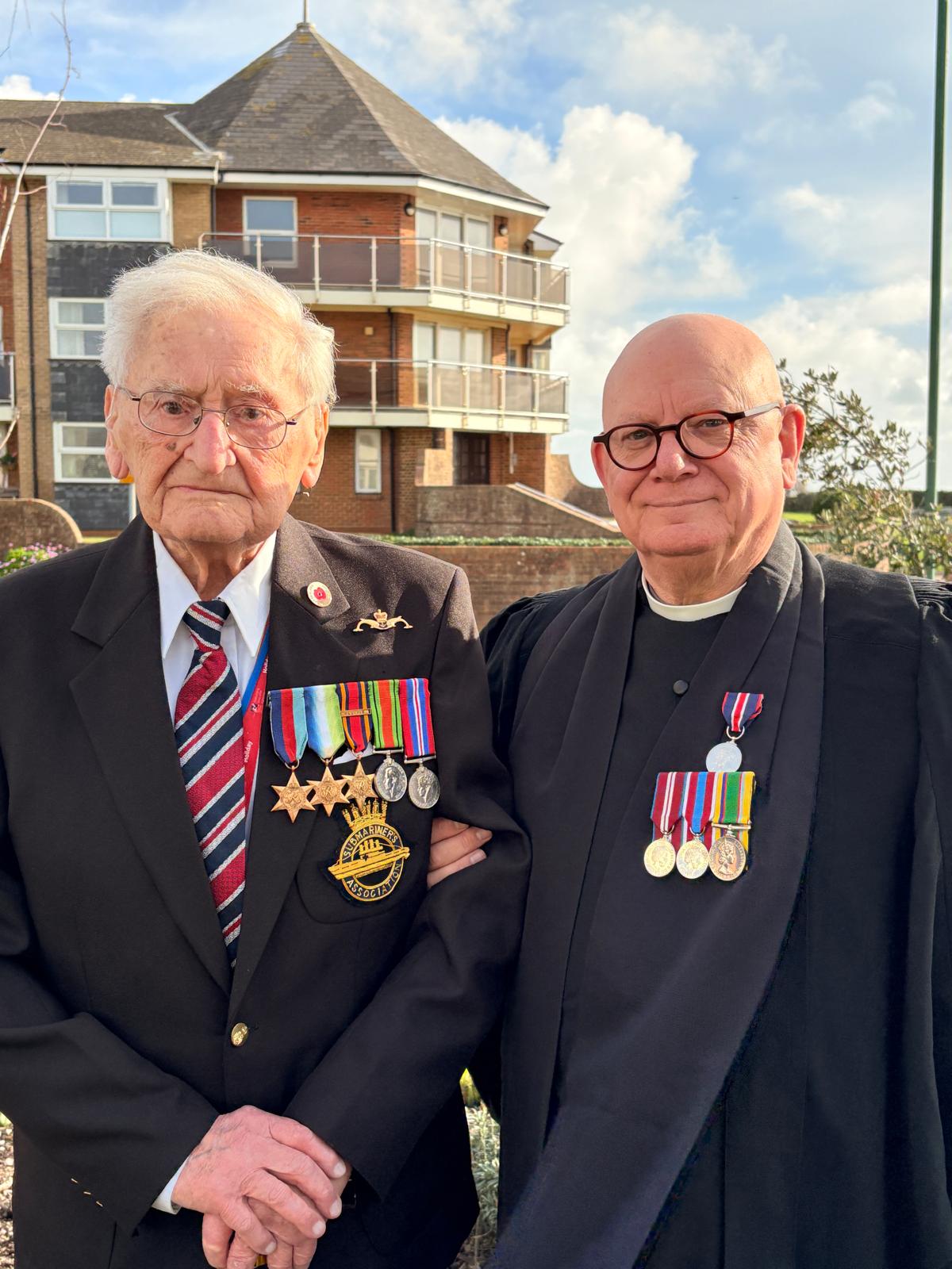 On the left, a WWII veteran with medals on his lapel stands arm in arm with a man dressed in a robe