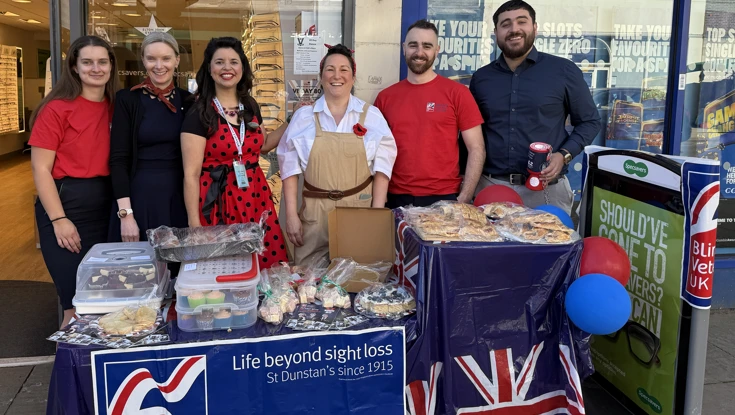 Six members of Specsavers staff (some dressed in 1940s style clothes) outside their store in Dorking with a table which is covered in a Union Jack table cloth and full of cakes. Also attached to the table is a Blind Veterans UK banner