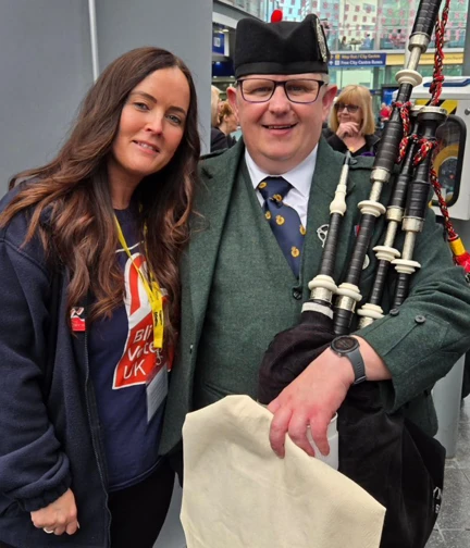 A Blind Veterans UK staff member stands next to a man in uniform holding bagpipes.