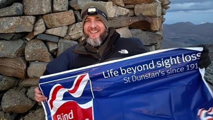 Benjamin with a Blind Veterans UK banner at the top of Scafell Pike with very little daylight.
