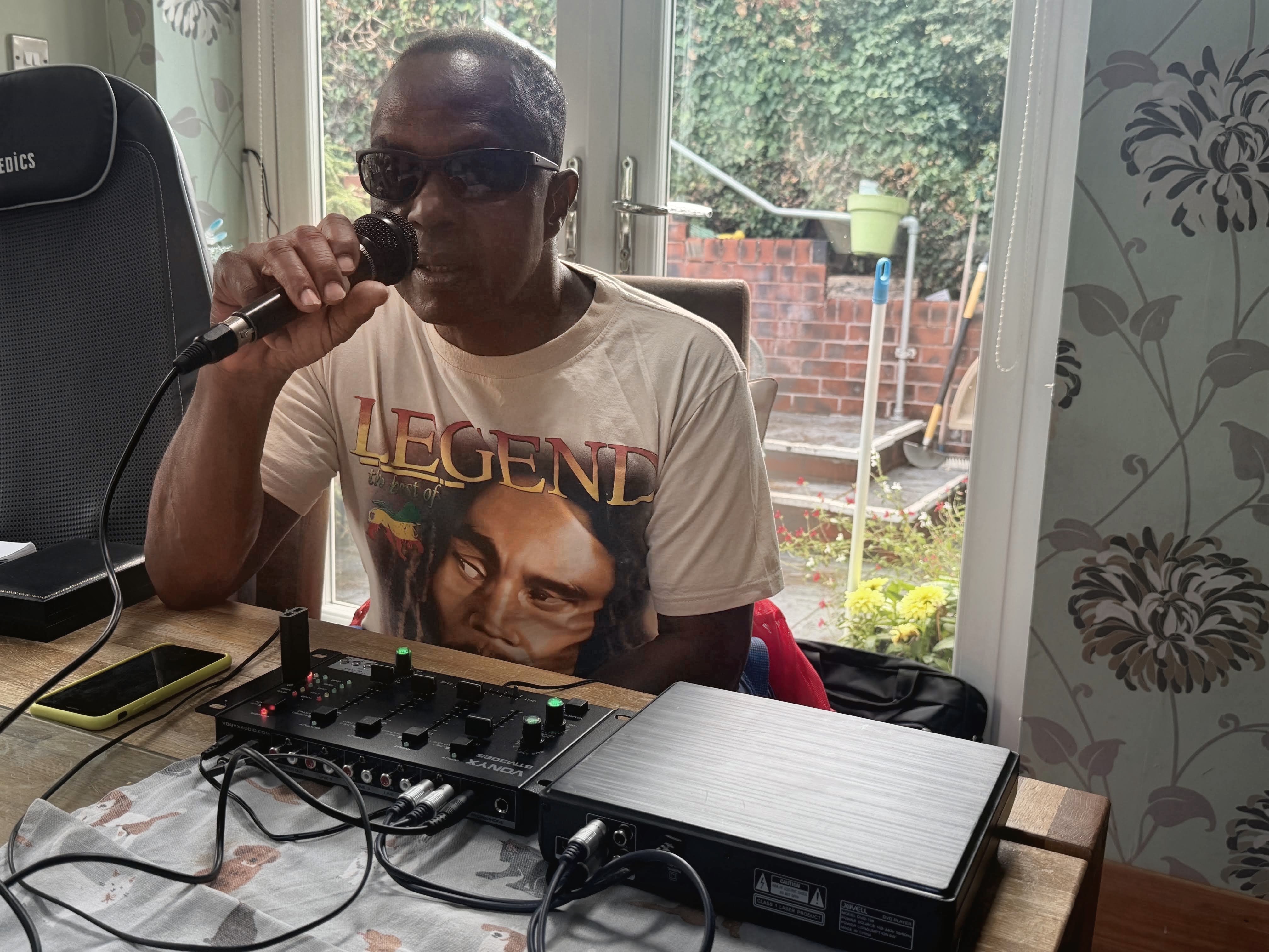 Michael speaks into a microphone and sits behind a desk with production equipment. He wears a Bob Marley t-shirt saying 'Legend'