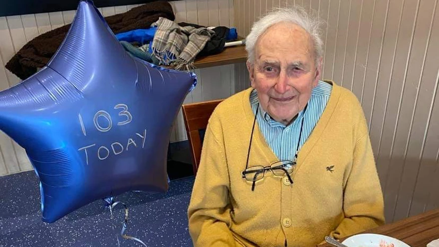 John looks into the camera with an finished desert plate in front of him and a balloon saying '103 today' next to him