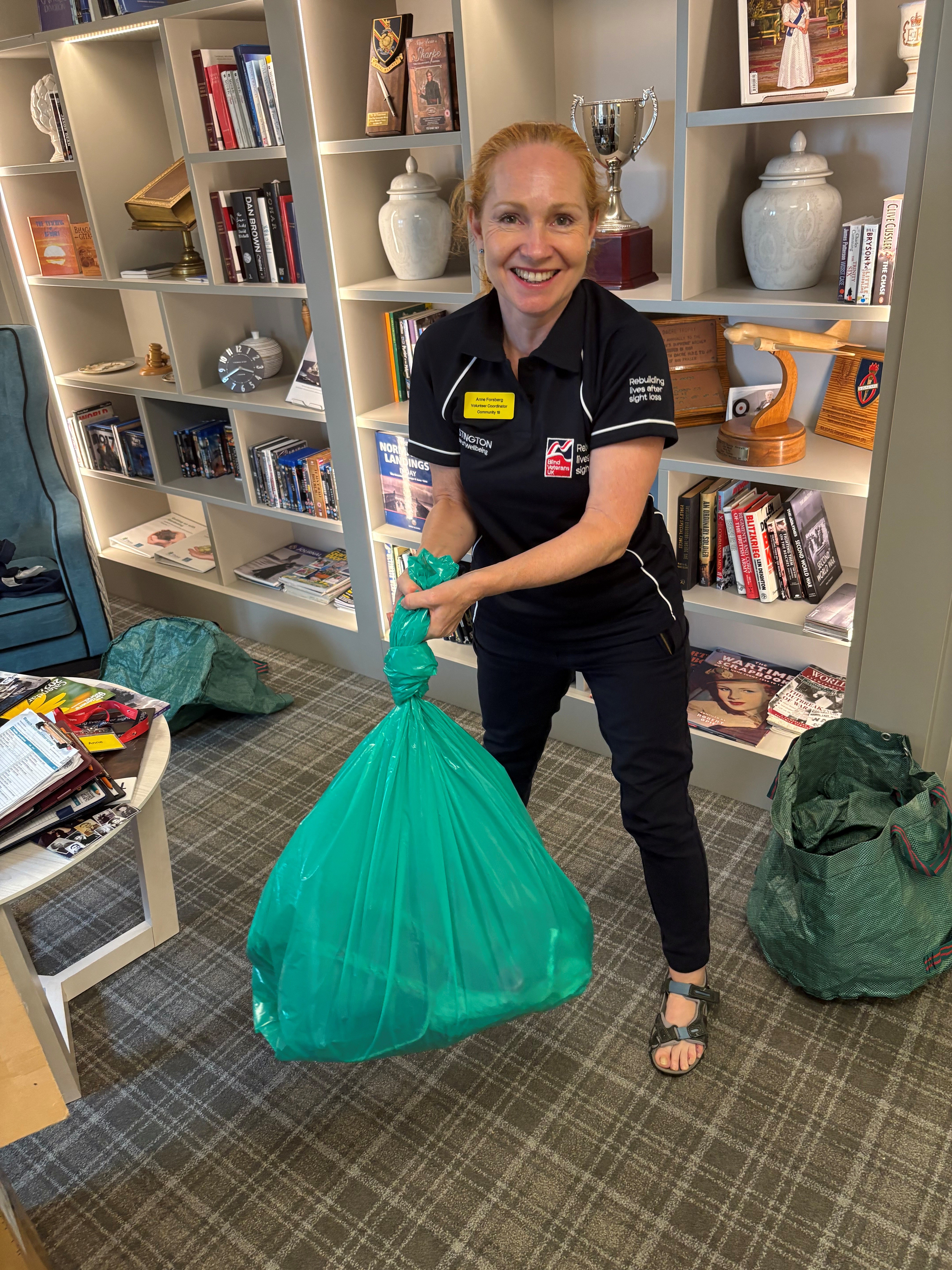 A lady with long red hair, wearing a navy blue t-shirt and trousers branded with the Blind Veterans UK logo stands looking at the camera and smiling. She is holding a yellow bin bag that is full of rubbish and tied in a knot at the top.