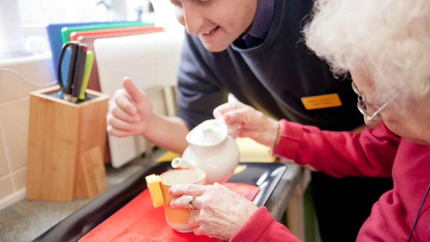 A blind veteran pouring a cup of tea, using a liquid level indicator, and a support worker leaning over her shoulder to help.
