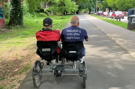 A volunteer and a blind veteran sat side by side on a tandem bike with four wheels cycling along a wide pathway.