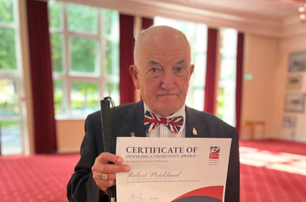 Bob is stood inside the Officer's Mess at HMS Sultan holding his Founder's Day award and white cane in one hand and a commemorative coin in the other.