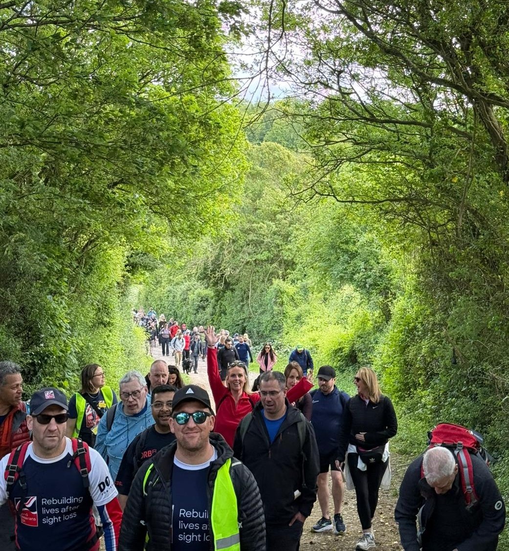 A large group of Operation Enigma participants walking uphill along a tree-lined path, with several people wearing Blind Veterans UK tops.