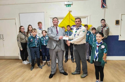 Clive and Cub leader Dan shaking hands and holding up cheque. They are surrounded by a group of cub scouts