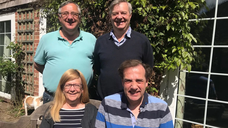 Linda and Nigel are sat on a wooden bench in the sun and Andy and Jerry are stood behind them
