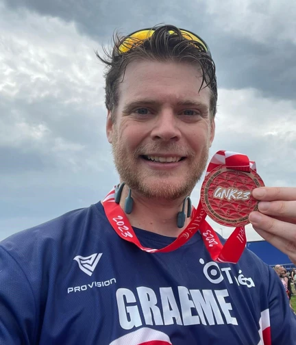 A flushed but happy looking Graeme holds his medal up as he smiles for the camera.