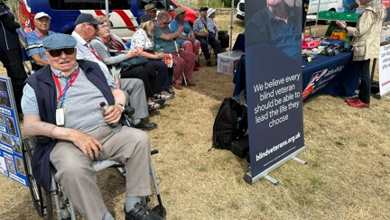 A group of blind veterans sitting on chairs in a field, next to a Blind Veterans UK stall