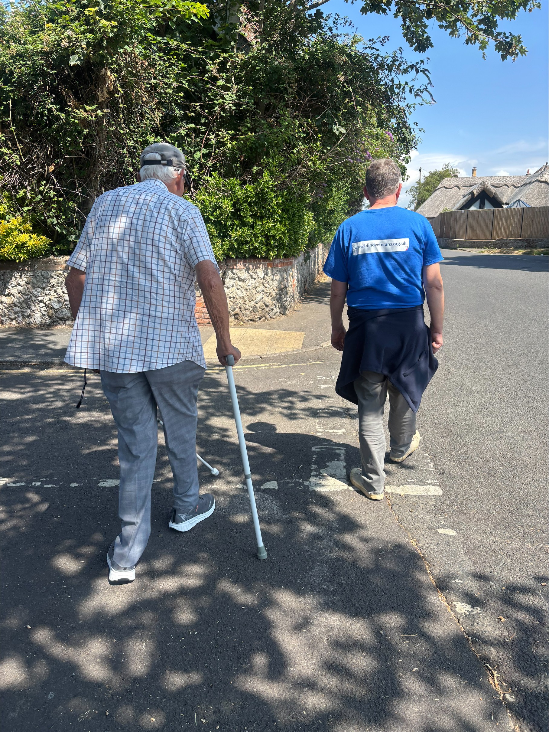 A photo of Richard taken from behind as he walks outside with a walking stick in one hand and white cane in the other