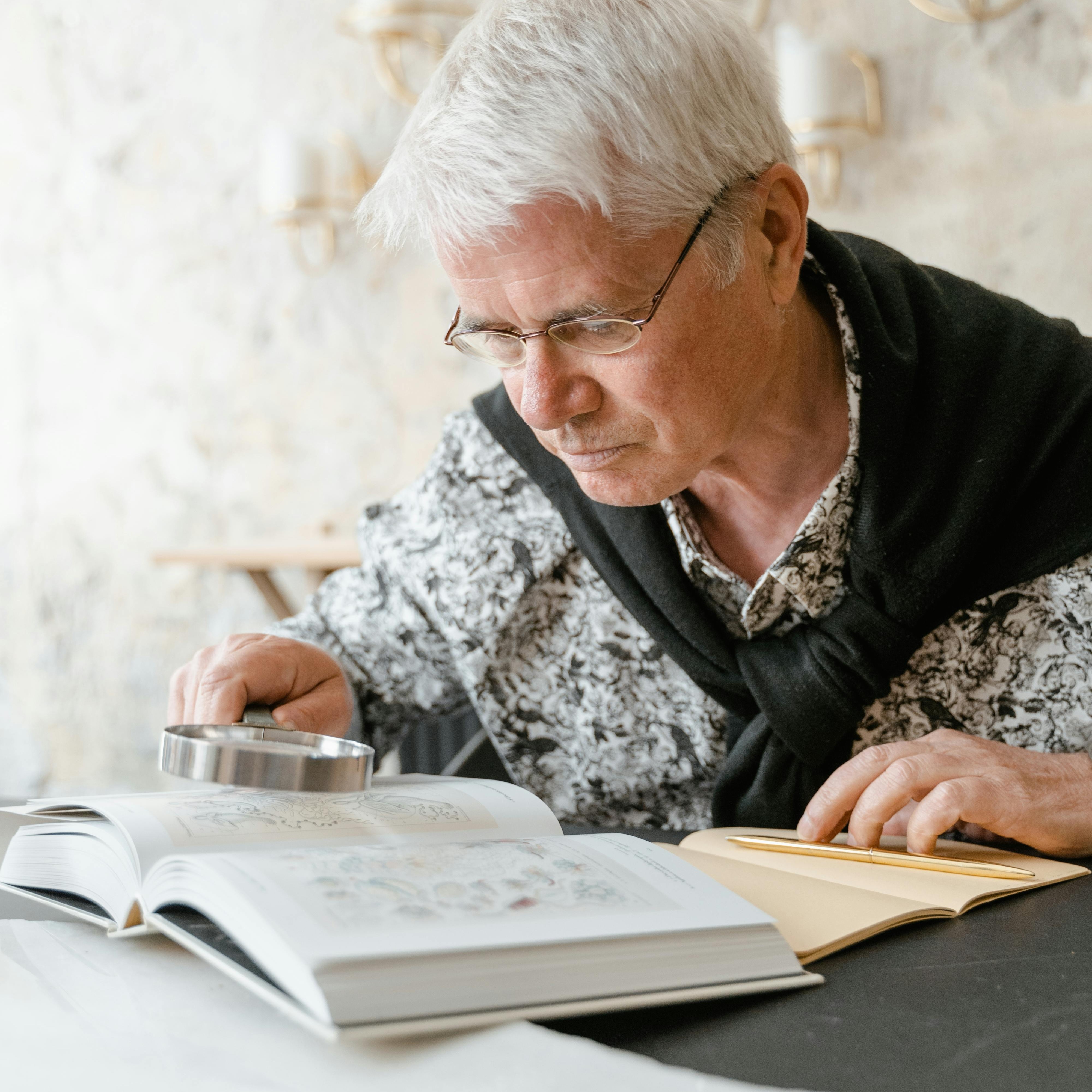 Close-up of a person wearing glasses using a handheld magnifying glass to read text in an open book, with a notebook nearby on a table.