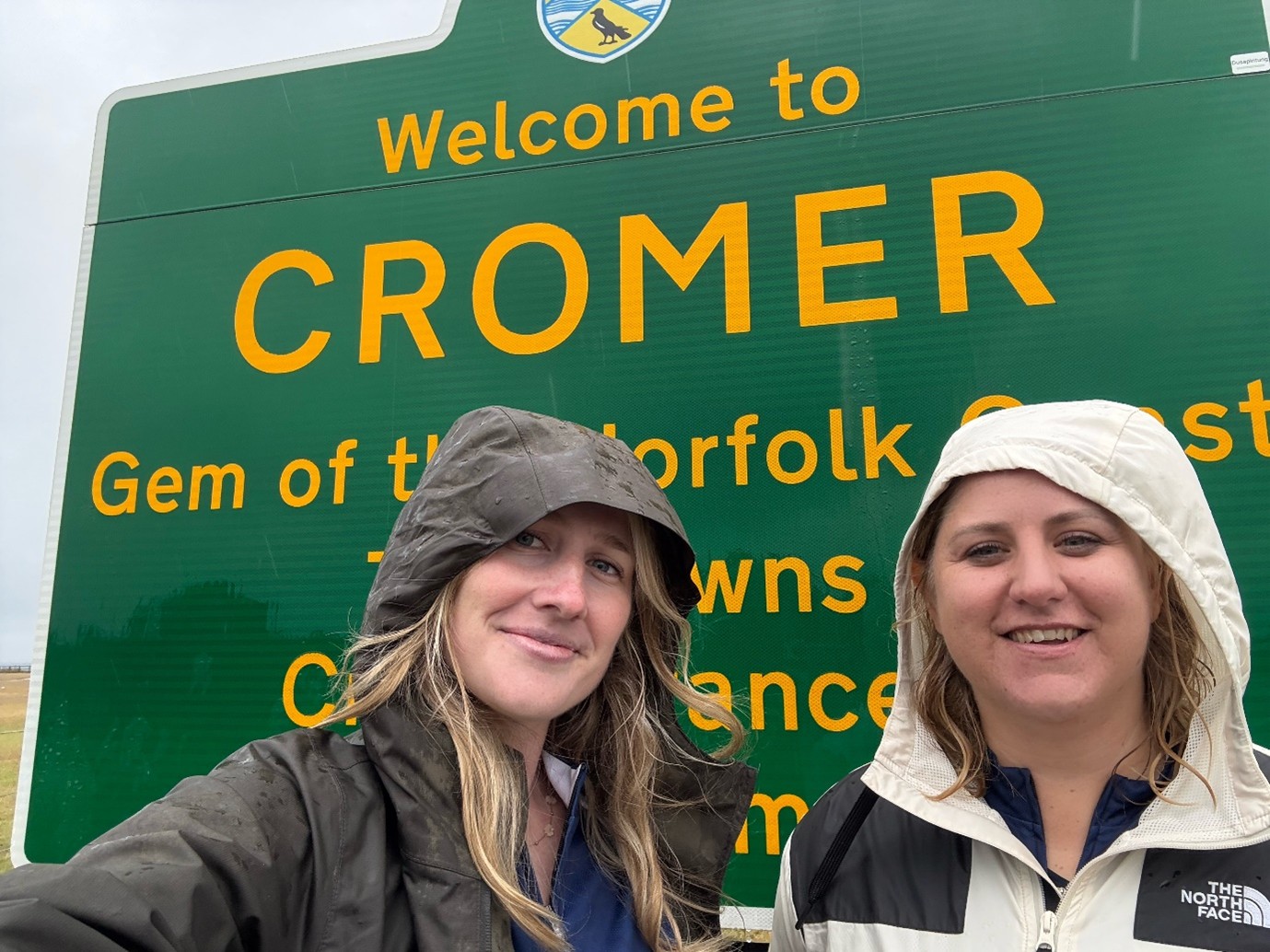 Kat and Rosie with their raincoats on and hoods up looking very wet. They stand in front of the Welcome to Cromer sign