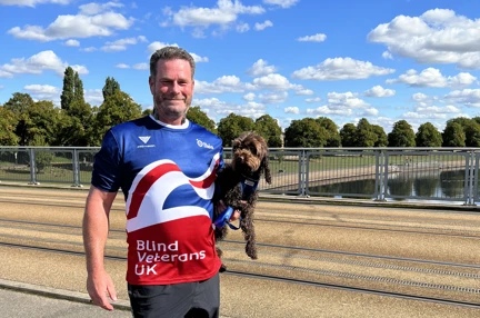 Simon stood in front of a canal in the sunshine wearing his Blind Veterans UK top and holding his dog