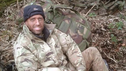 A young Simon sitting on the ground in a wooded area wearing camouflage clothing
