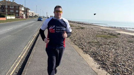 Wayne wearing his Blind Veterans UK t-shirt and tinted yellow glasses running alongside the pebble beach