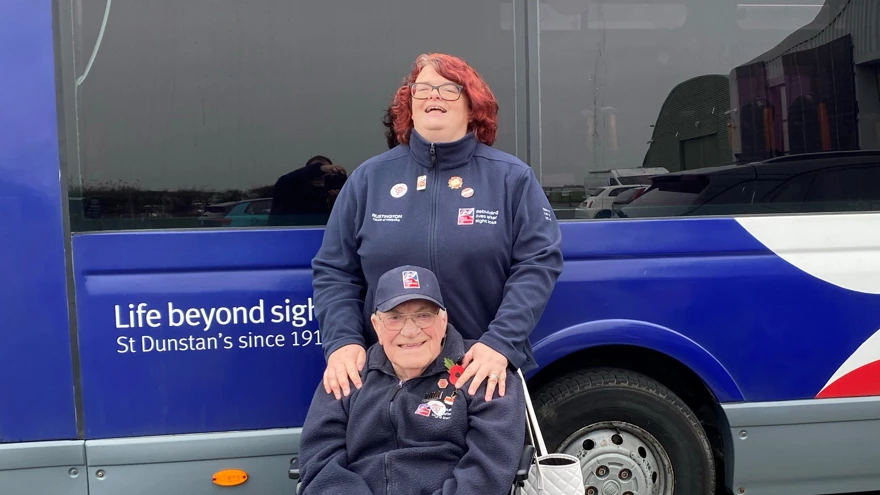 Paula stood behind blind veteran Bill. Bill is sat in his wheelchair and Paula has he hands on his shoulders. Both are smiling for the camera.