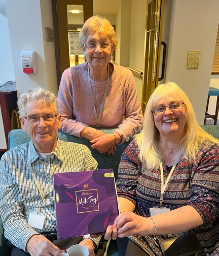 Three people sitting on a sofa, holding a box of Cadbury's milk tray chocolate and smiling