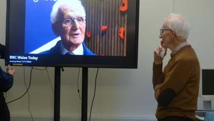 A man standing in front of television screen showing a picture of him