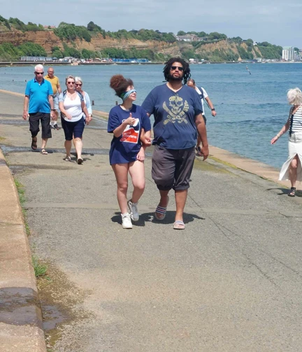 A lady in a Blind Veterans UK t-shirt is blindfolded as she walks along a promenade with the sea alongside her. She has her arm linked with a man who is guiding her