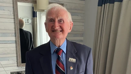 A man stands smiling at the camera. He is wearing a navy blazer and a navy, red and grey striped tie.