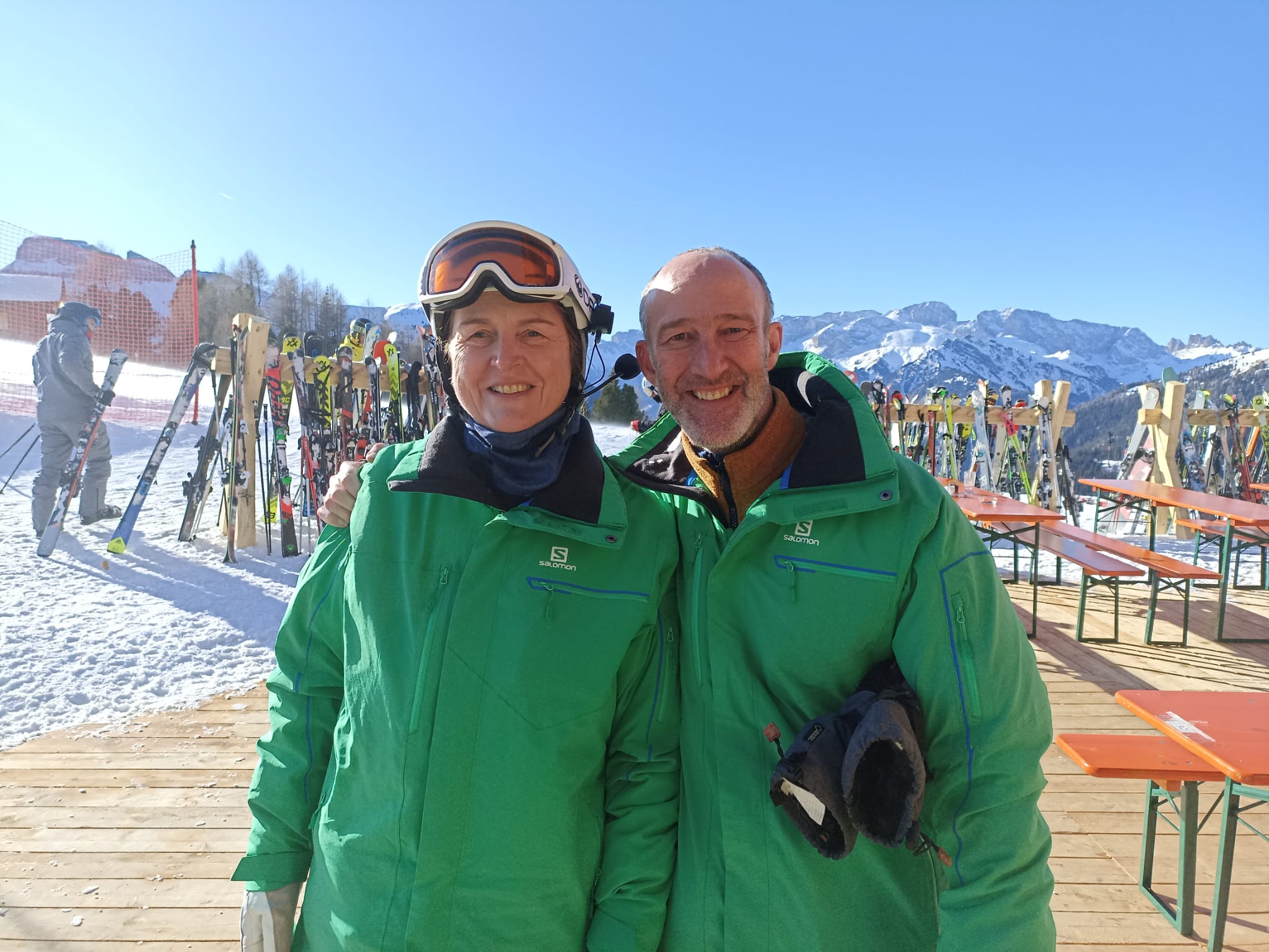 Blind veteran Jan (left) and volunteer guide Bob (right) smile together at a snowy ski resort. Both wear bright green jackets, and Jan wears a ski helmet and goggles. Skis and snow-covered mountains are visible behind them.
