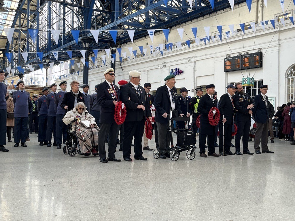 A group of people wearing formal clothing, berets, poppies and military medals stand inside a railway station, several holding red poppy wreaths or using mobility aids. A group of RAF Air Cadets stands behind them.