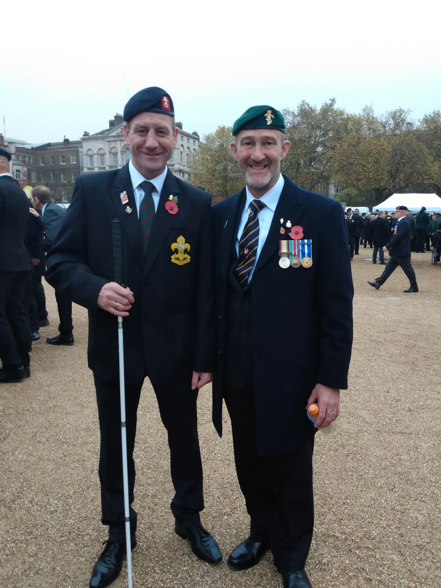 Two people wearing dark formal clothing, berets, poppies and military medals stand together at a Remembrance event. The person on the left holds a white cane.