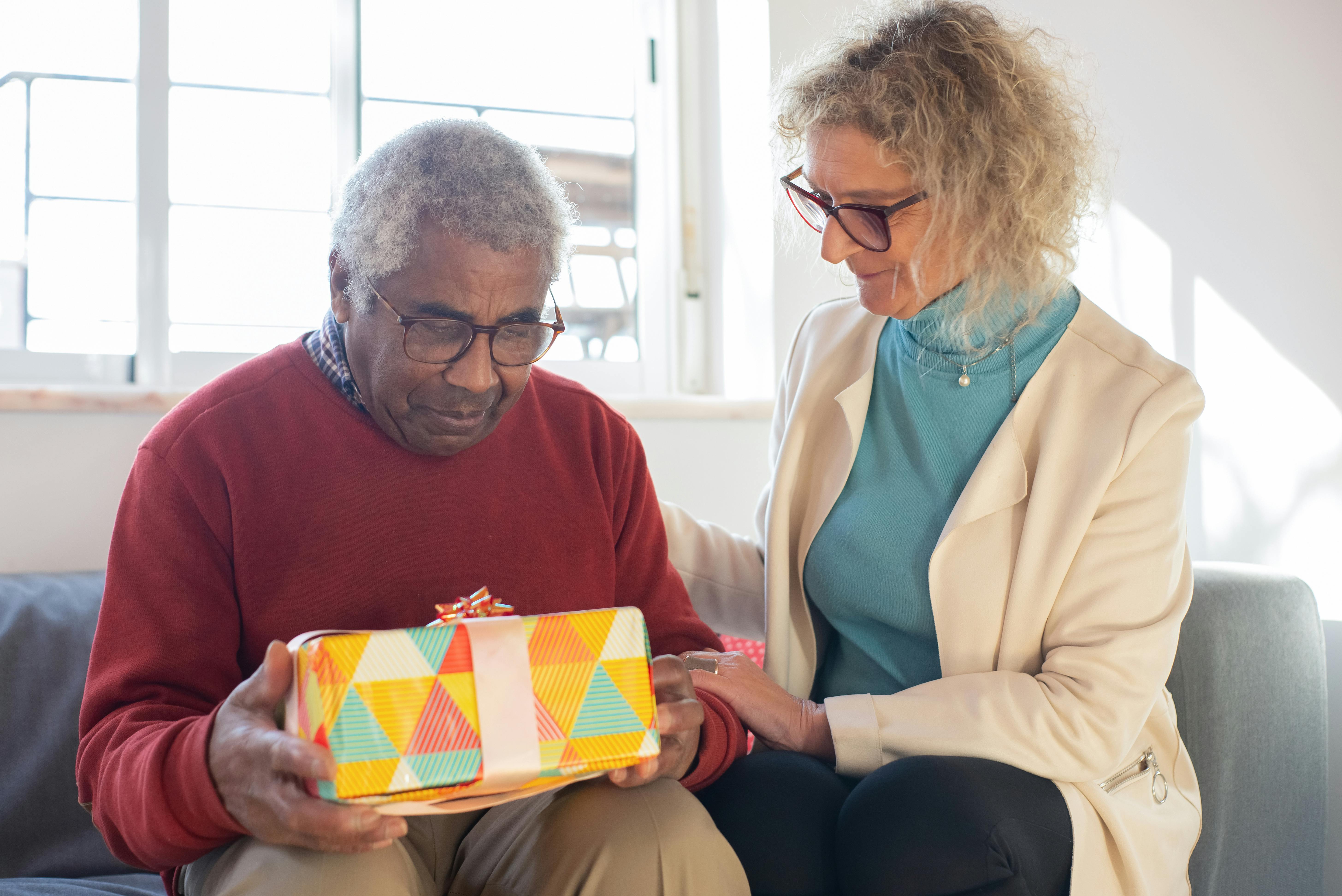 Two people sitting on a sofa indoors; one person holds a colourful wrapped gift while the other places a hand on their arm, with a bright window in the background.