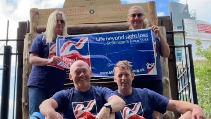 Janine and three others posing with the sign that signals they have completed the length of Hadrian's Wall. They are all in their Blind Veterans UK t-shirts and holding up a Blind Veterans UK flag
