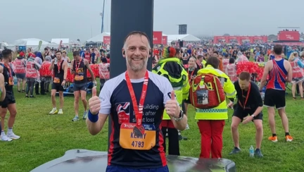 Kenny wearing his Blind Veterans UK running top with his medal around his neck and both thumbs up. Lots of runners in the background