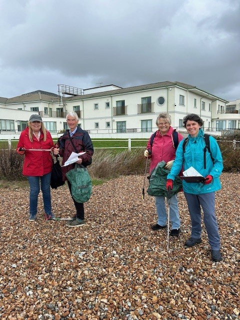 A group of four people wearing waterproof coats stand in a line on a pebble beach. They are looking at the camera and holding litter pickers and bags.