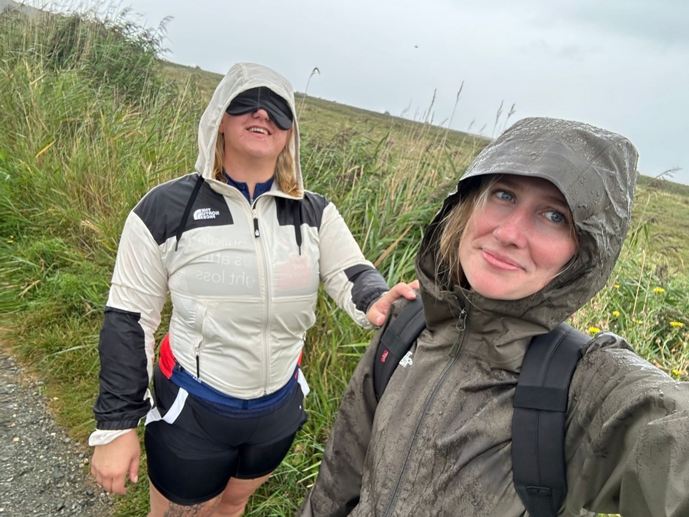 Kat is blindfolded and had her hand on Rosie's shoulder as they stop to pose for a photo. Both women are wearing rain coats with their hoods up and it is raining