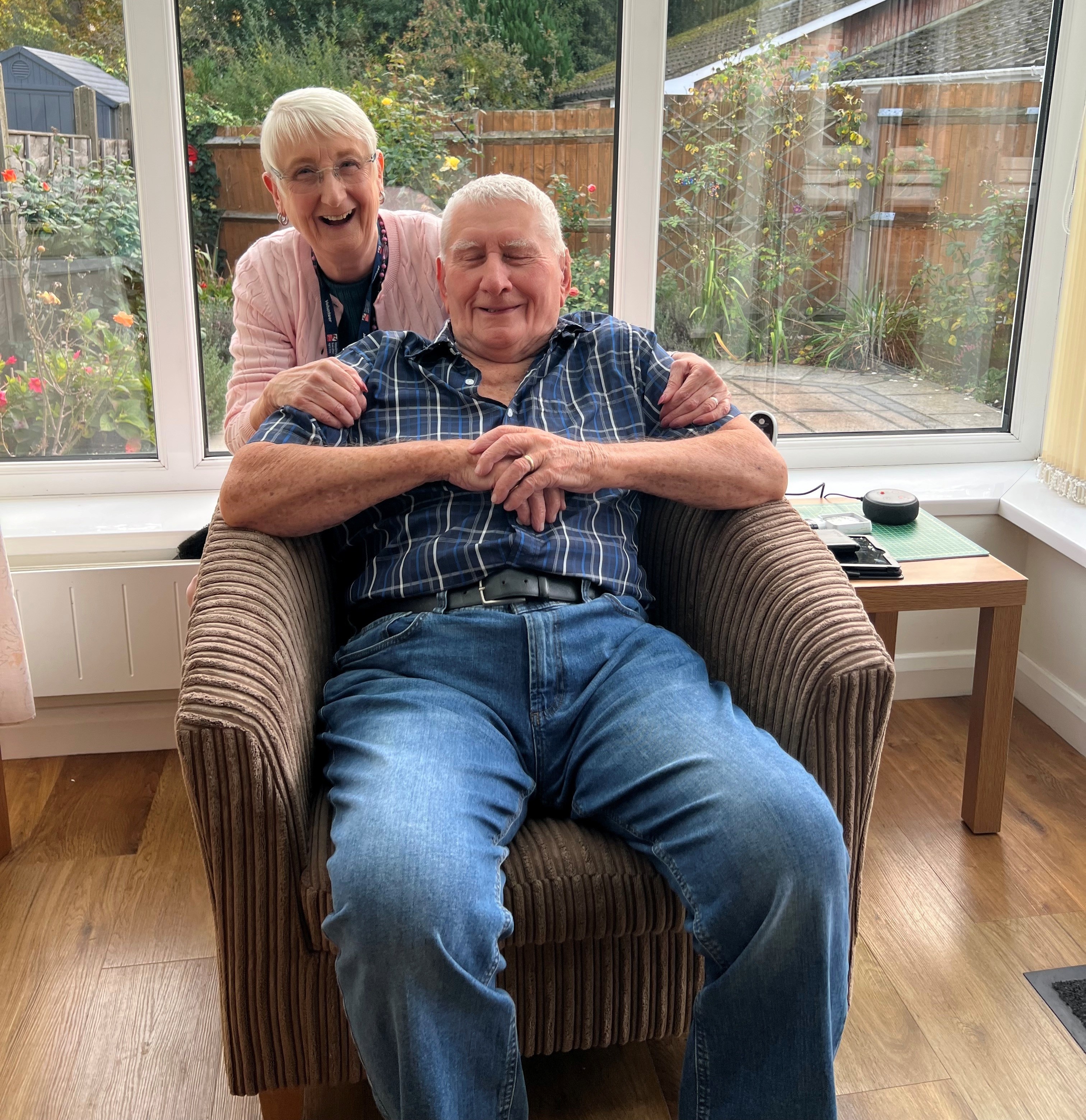 A man with grey hair and wearing a blue and green checked shirt and jeans is sitting in an armchair looking at the camera and smiling. A woman with short grey hair wearing glasses, a pink cardigan and black top is standing behind him on the left of the picture. She is leaning over the chair with her hands on his shoulders, looking at the camera and laughing.
