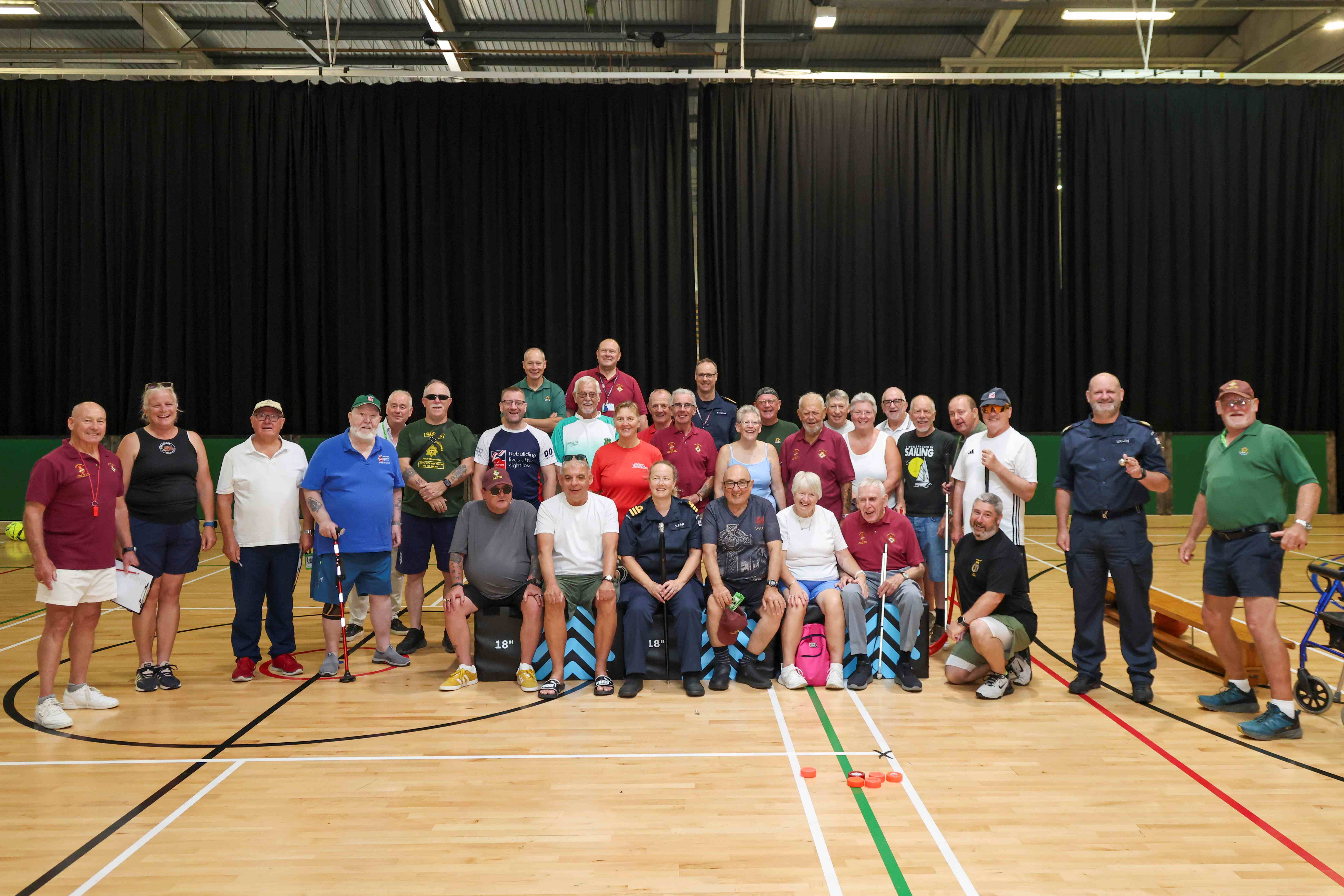 The group sit lined up on chairs in a sports hall and look at the camera