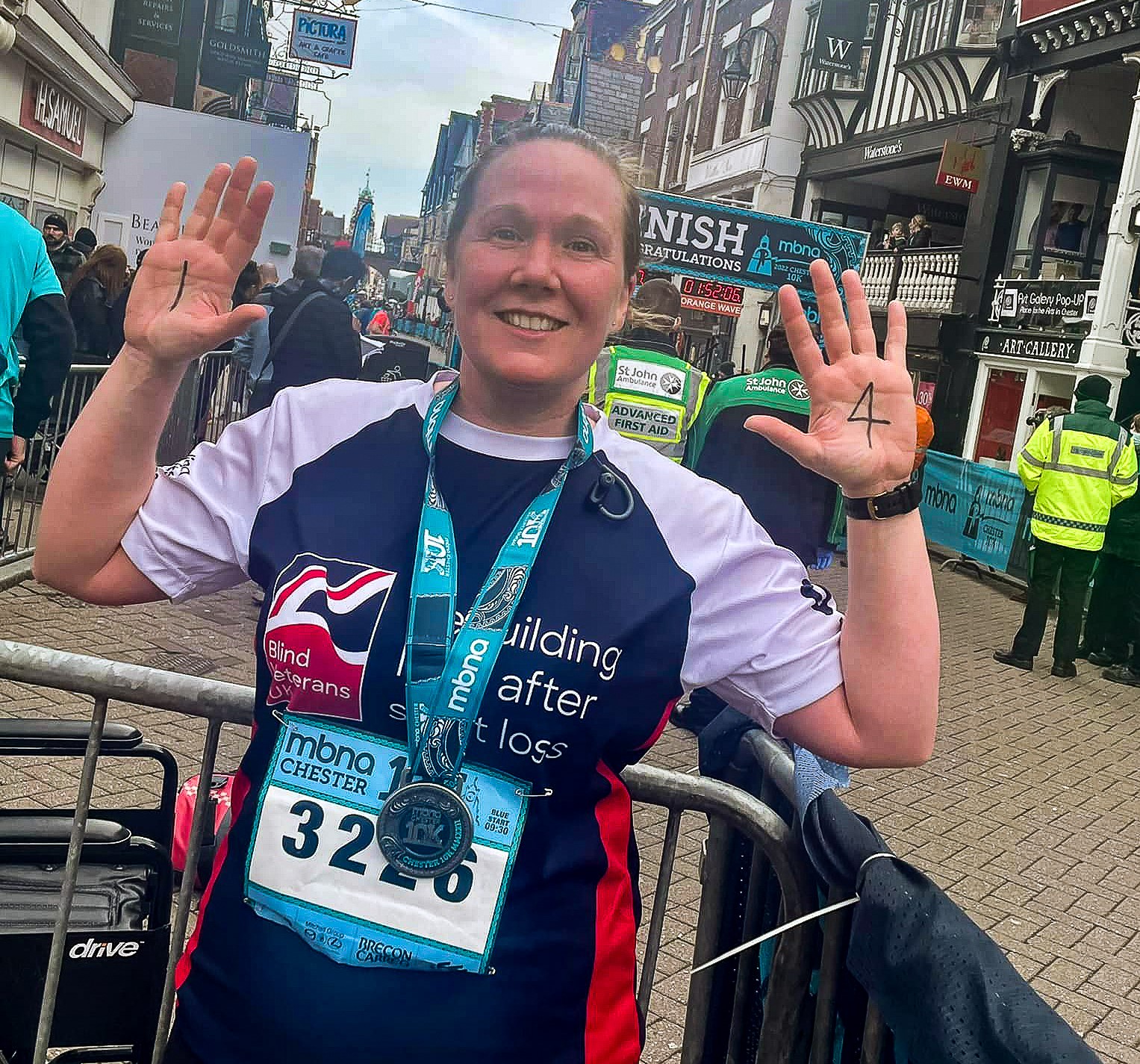 Supporter Alison wearing her marathon medal and Blind Veterans UK training top, holding up her hands to show her palms, with the number 1 written on her left palm, and 4 on her right palm.