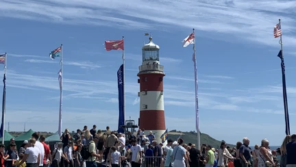 Crowds gathered near a lighthouse and multiple flying flags at Plymouth Armed Forces Day,
