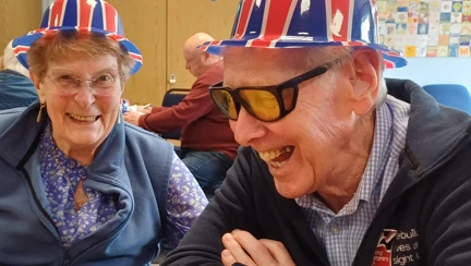 Two blind veterans smiling and wearing Union Jack hats, enjoying a VE Day celebration at a decorated venue.