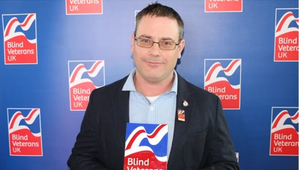 Blind veteran Andy is wearing a suit and holding his Founder's Day award. He is looking into camera and there is a Blind Veterans UK backdrop.