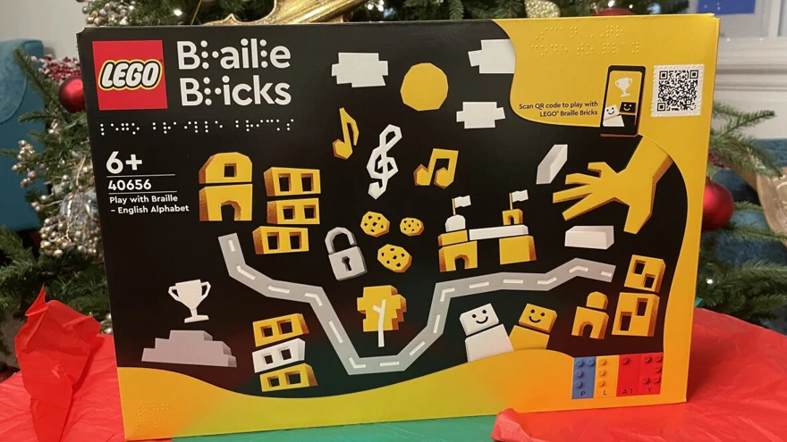 A box of Braille lego bricks, sitting on top of some festive coloured paper hats with a Christmas tree in the background