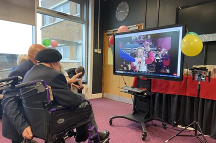 Blind veteran Ken in front of a TV screen with Margaret waving from the video call