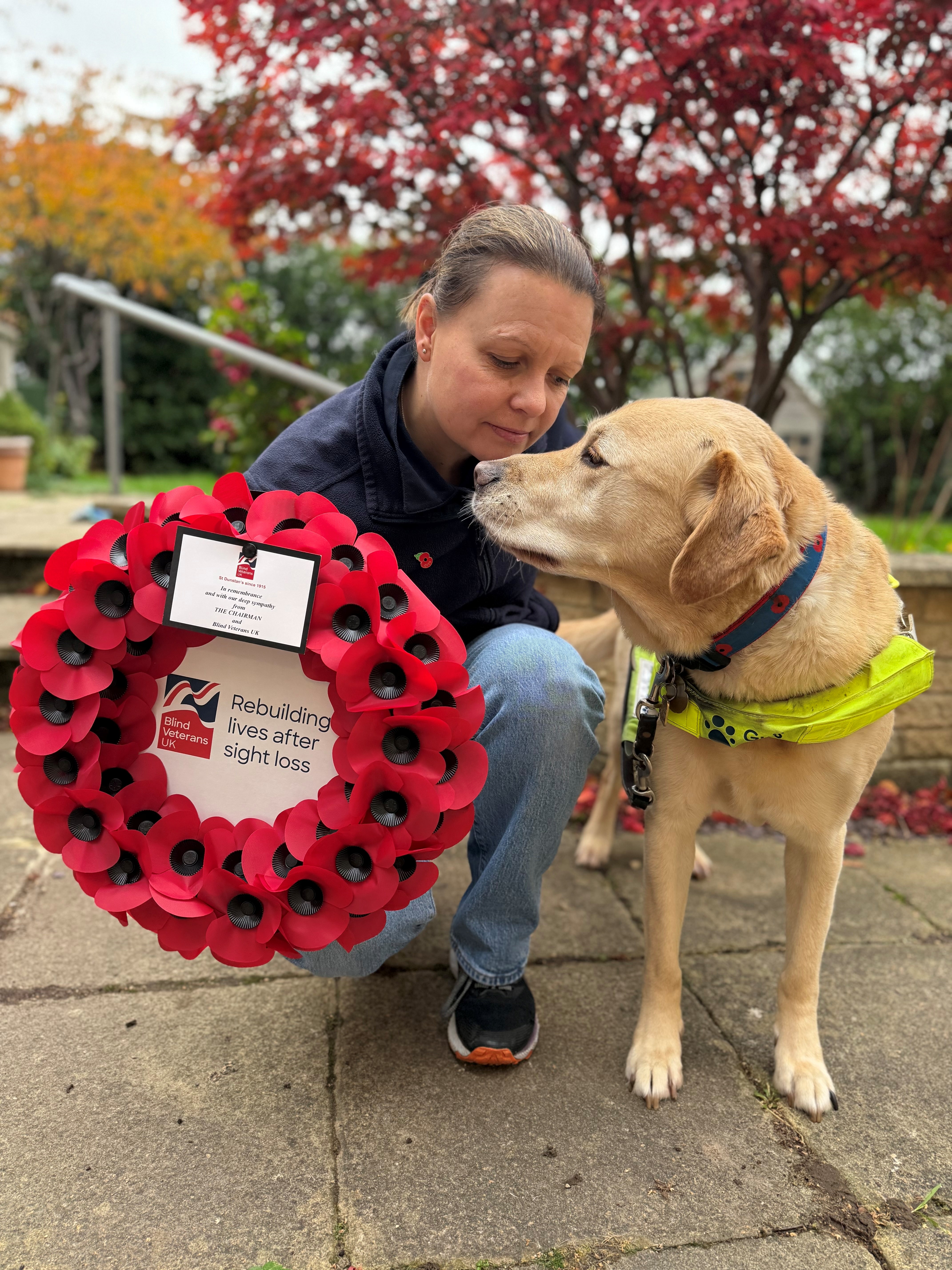 Kelly is kneeling on the ground in her garden with her guide dog Archie, a golden lab cross retriever, beside her. Archie has his harness on and a collar with poppies. Kelly is holding a wreath.