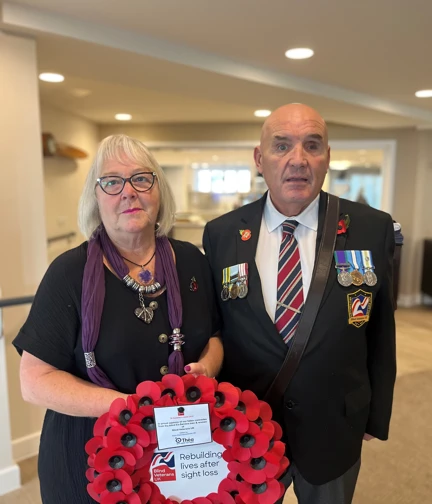 Alan and Shirley are stood side by side. Alan is wearing his medals and Shirley is holding a wreath which has been gifted by Thea Pharmaceuticals