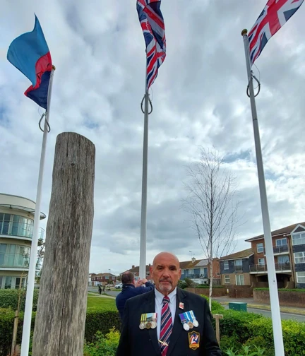 A blind veteran in uniform with medals stands near a flagpole at the Rustington Centre on VE Day.