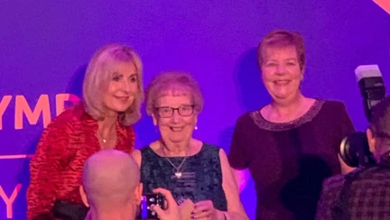 Gill is standing with the presenter and posing for photos by the photographers