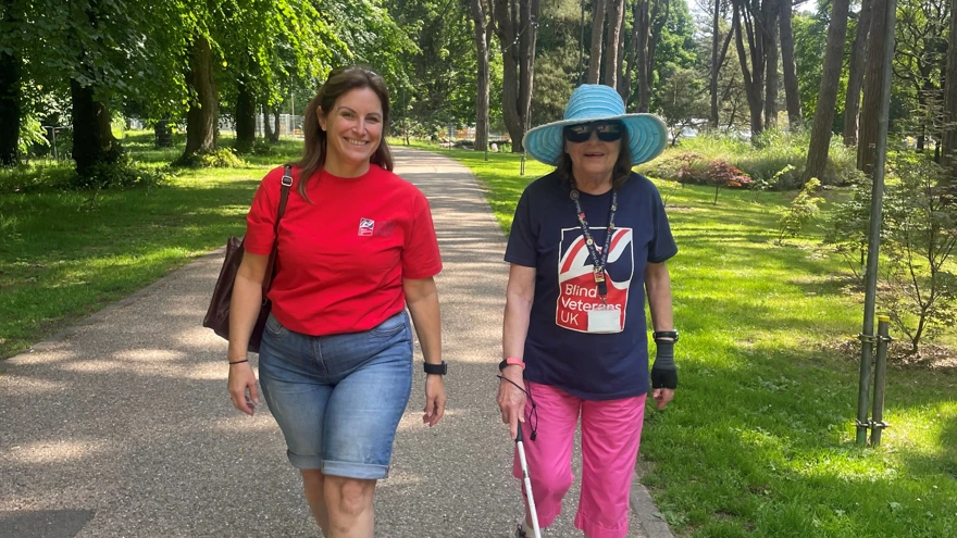 Beth and Noeline walk side by side along a tree lined pathway. Noeline is wearing a Blind Veterans UK t-shirt and using her cane.