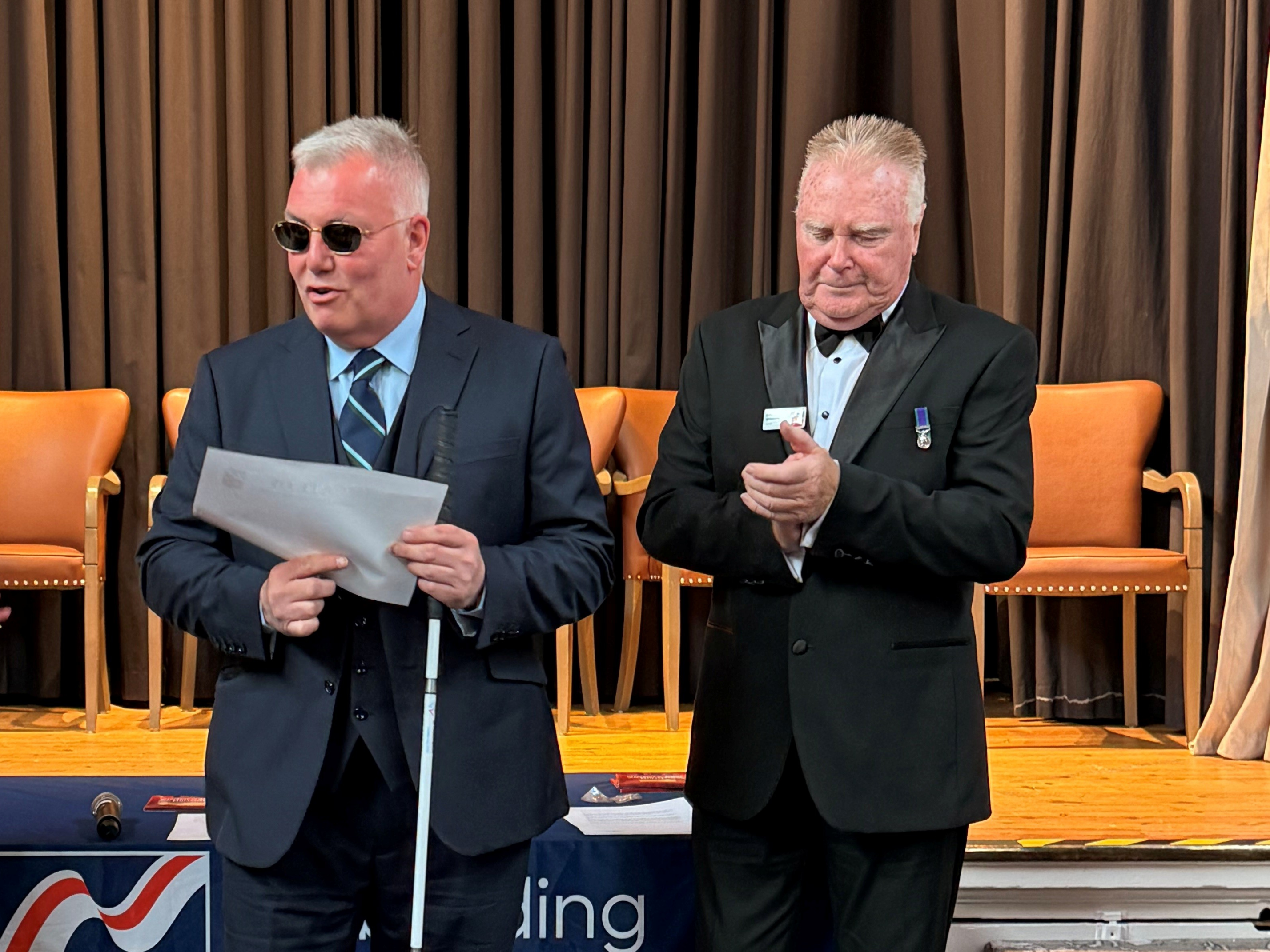 Blind veteran Niall is wearing dark sunglasses and holding a white cane as he receives his award. President Colin Williamson stands next to him clapping. 