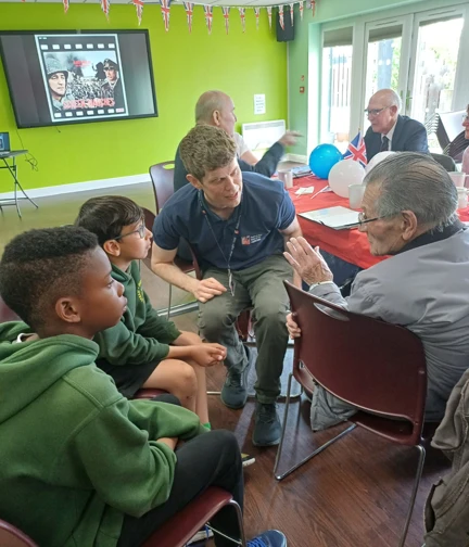 Blind veteran Ted sits in a classroom speaking to school children and a Blind Veterans UK staff member, sharing his experiences.
